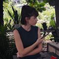 The picture shows a white woman with black hair looking to the side. She is wearing a black tank top and there are plants behind her. 