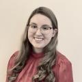 The image is a headshot of a young white woman with brown hair wearing a red shirt and glasses. She is smiling 