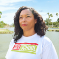 The picture shows a Latina woman with curly black hair and red lipstick smiling at something in the distance. She is standing in front of a body of water. 