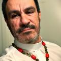 A white man with short black hair is looking at the camera, wearing a white priest's robe and a rosary