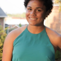 The photo shows a Black woman with short hair smiling at the camera. She is wearing a dark teal tank top. 