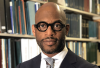 The picture shows a bald Black man with facial hair wearing black glasses in front of a bookshelf, wearing a suit. 