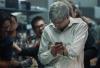 A photo from the docufilm ‘Blackberry.’ Actor Jay Baruchel is Mike Lazaridis, co-CEO of Blackberry. He has short gray hair, glasses, and wears a white dress shirt. He glares down at a phone with wires plugged into it. People behind him are cheering.