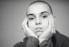 A black-and-white photograph of Sinéad O’Connor in ‘Nothing Compares.' Her head is shaved and she is wearing a long-sleeve shirt. She is resting her head in both of her palms with her fingers clasped over both cheeks. 