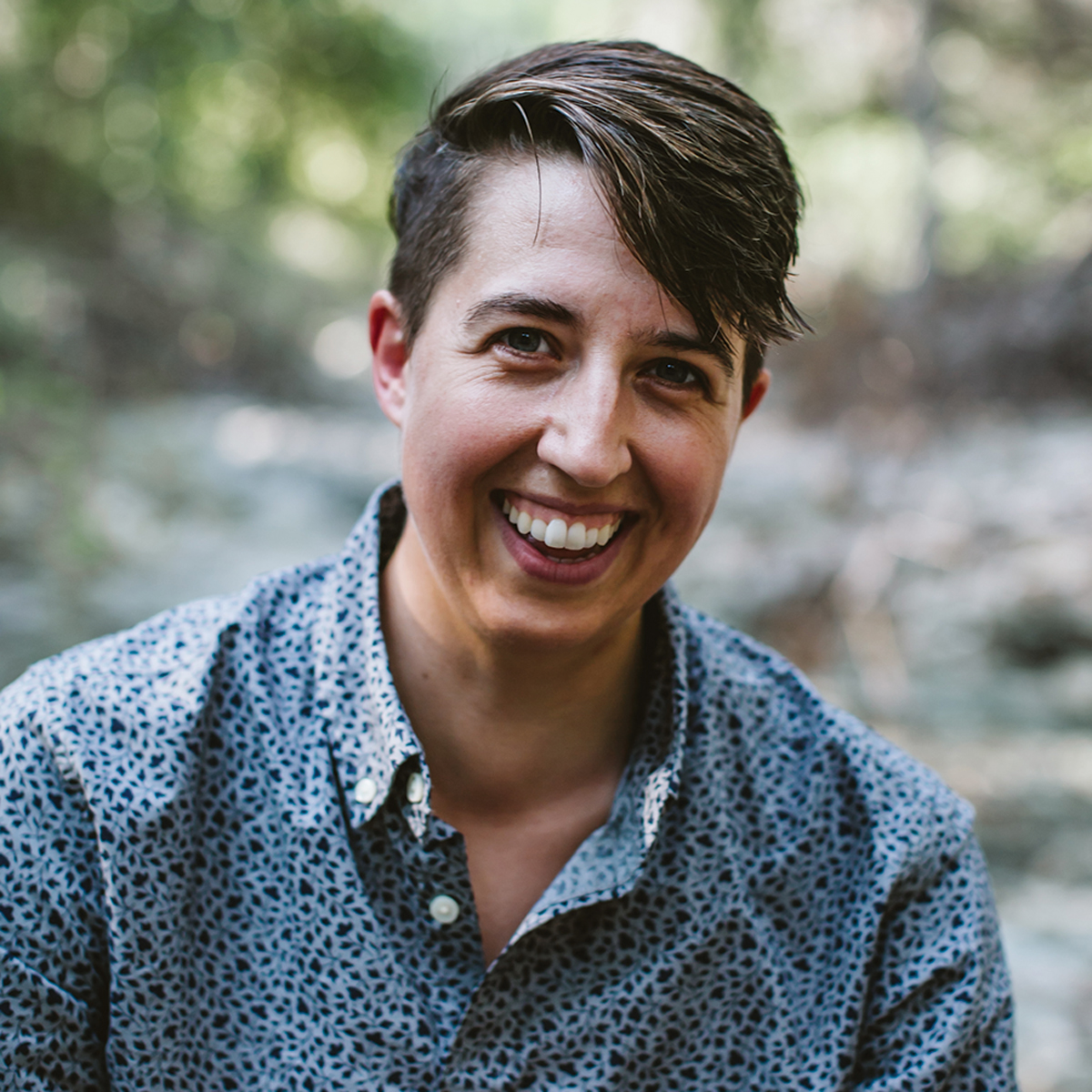 A photo of Abi Robins: author of 'The Conscious Enneagram.' They are a white non-binary trans person with short dark blonde hair. They are smiling and wearing a light blue shirt with various shades of blue dots.