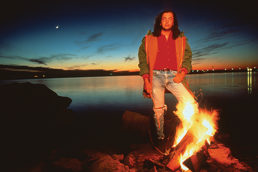 A photo of Rich Mullins with his back to a lake at sunset. He's standing in front of a campfire with the glow illuminating his red shirt, green and orange jacket, and blue jeans. He has long brown hair and is holding a stick.