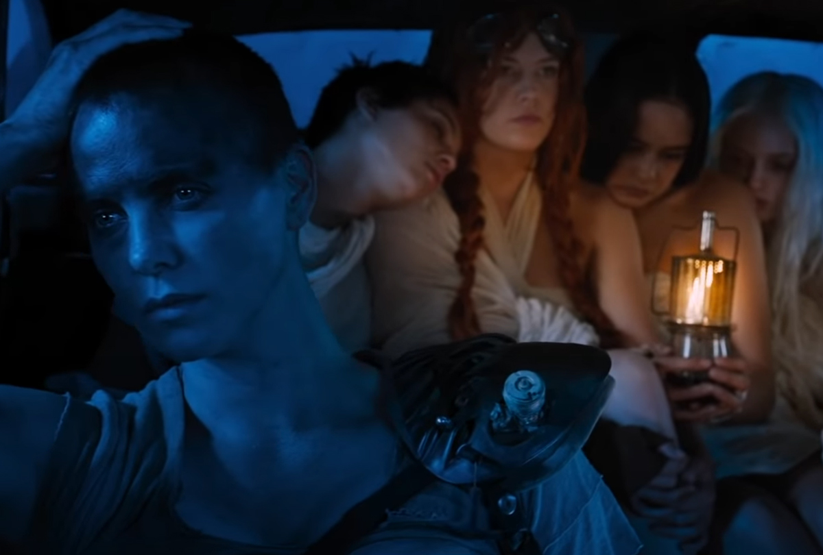 Furiosa (actress Charlize Theron) in 'Mad Max: Fury Road' sits in the passenger seat of a cramped car. Four women are huddled together in the backseat, holding a lamp that illuminates the cool blue night.